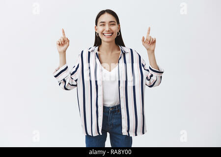 Ritratto di bellezze naturali ed eleganti donna in jeans e camicia a righe, sollevando le mani e rivolto verso l'alto con le dita indice, sorridente gioiosamente alla telecamera, essendo divertito e intrattenuto oltre il muro grigio Foto Stock