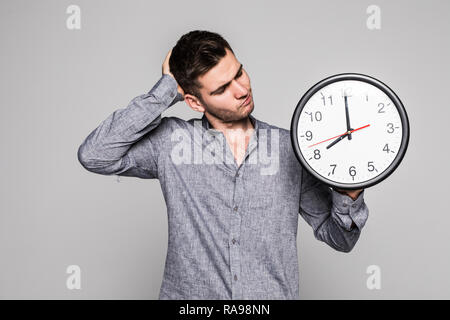 Ritratto di un sorridente uomo informale triste perchè tardi holding orologio da parete su sfondo grigio Foto Stock