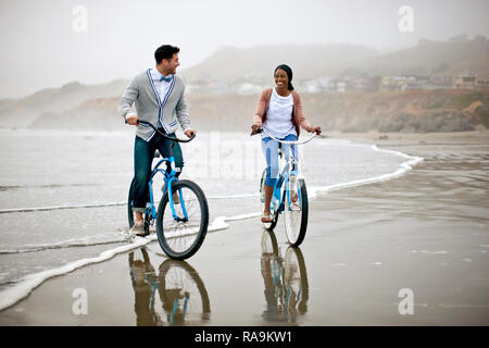 Felice coppia giovane la bicicletta lungo una spiaggia con la bassa marea. Foto Stock