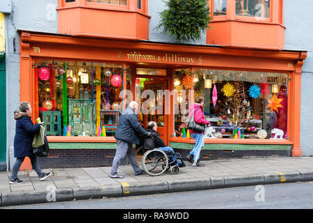 La persona disabile che viene spinto in una sedia a rotelle di fronte ad una nuova epoca shop, Glastonbury, Somerset, Regno Unito - Giovanni Gollop Foto Stock