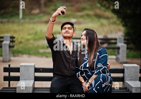 Indian paio poste all'aperto, seduto su un banco di lavoro insieme e guardando il telefono cellulare, rendendo selfie. Foto Stock