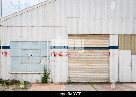 Un vecchio bianco parete corrugata con rosso e blu strisce sbiadite, rullo porta e finestrini rotti Foto Stock