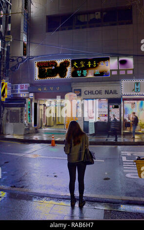 Una con i capelli lunghi donna stand, torna alla fotocamera, stagliano sul bordo di una strada di città inondate di luce al neon di Seoul, Corea del Sud Foto Stock