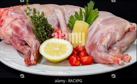 Materie carne di coniglio con erbe e spezie su una piastra bianca su sfondo nero preparato per cucinare. Foto Stock