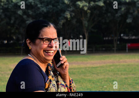 Senior donna indiana di ridere e di parlare al telefono, seduti in un parco a Delhi, India Foto Stock