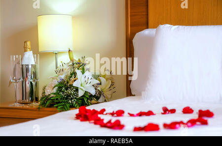 Accogliente e romantico letto con petali di rosa con focus sul comodino. Biancheria da letto bianca, fiori, champagne flauti, luna di miele impostato Foto Stock