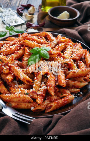 Italiano arrabiata penne su una piastra di argilla con basilico fresco e grattugiato formaggio di capra su un tavolo rustico con panno marrone, verticale vista da sopra Foto Stock