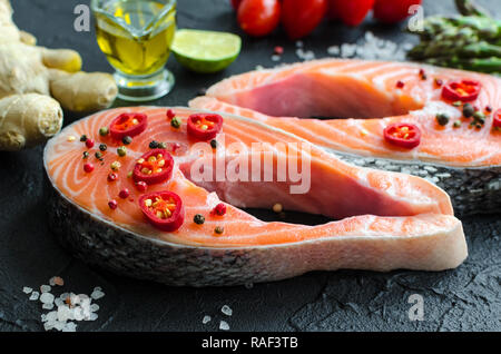 Due crudo fresco filetti di salmone con spezie e verdure: asparagi pomodori, lo zenzero, i calli di pepe, sale e pepe, peperoncino, limone e olio di oliva su nero ba Foto Stock