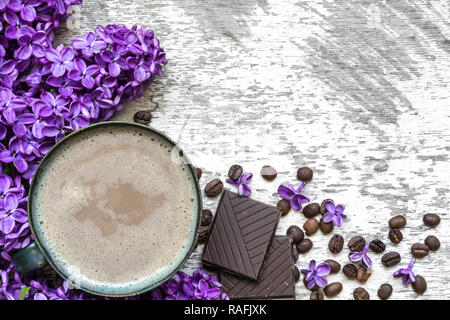 Tazza di caffè con i chicchi di caffè, cioccolato e fiori lilla bianco su sfondo di legno. Lay piatto. vista superiore Foto Stock