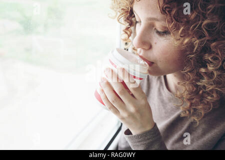 Close-up verticale di una bella e giovane donna con occhi blu e ricci capelli biondi beve caffè o tè mentre si è in viaggio Foto Stock