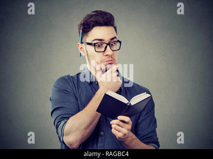 Giovane uomo intelligente in bicchieri libro di lettura con note interessanti mentre si tocca il mento nella contemplazione Foto Stock