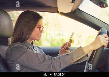 Vista laterale di distrarsi giovane donna in bicchieri utilizza lo smartphone durante la guida auto Foto Stock