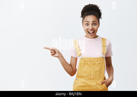 Un nuovo straordinario prodotto in magazzino. Attraente e gioiosa eccitato attraente americano africano femminile nel trendy tuta gialla, tenendo la mano in tasca, rivolta a sinistra e sorridente largamente, impressionato Foto Stock