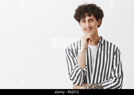 Creativo, elegante e hipster ragazzo con i baffi e ricci capelli scuri nel quartiere alla moda di camicia a righe tenendo la mano sul mento e sorridente dall interesse e curiosità essendo incuriosito da shop assistant Foto Stock