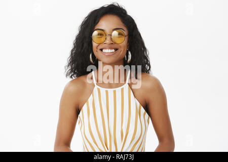 Vita-up shot di affascinanti di moda americano africano giovane femmina traveler in simpatici occhiali da sole giallo e striato top sorridente gioiosamente godendo i fine settimana su sfondo grigio Foto Stock