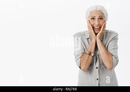 Studio shot di senior donna reagire a sorpresa. Ritratto di toccato e felici carino vecchia signora con i capelli bianchi toccando le guance divertito e sorridente broadlt ricevere regalo bellissimo oltre il muro grigio Foto Stock