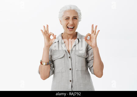 Vita-up shot del carismatico felice e carino awesome granny winking gioiosamente sorridente e mostra bene i gesti confermando ha affrontato tutto e noi non avere preoccupazioni su sfondo grigio Foto Stock