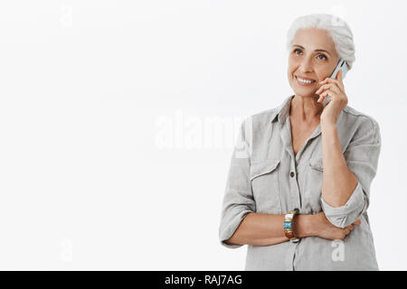 Vita-up shot di felice carismatico ed eccitato carino anziani femmina con i capelli bianchi in camicia casual e bracciale holding smartphone vicino all orecchio a parlare con un amico su sfondo grigio casualmente Foto Stock