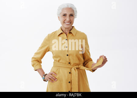 Cerca elegante come sempre. Ritratto di eccitato e carismatica donna anziana nel quartiere alla moda di rivestimento giallo giocando con la cinghia e sorridente gioiosamente cercando piuttosto e sensazione di giovani su sfondo grigio Foto Stock