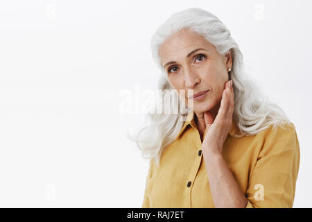 Fiducioso e elegante femminile donna anziana con lunghi capelli bianchi in giallo elegante trench coat toccando delicatamente la faccia e guardando la telecamera soddisfatto di come la pelle apparirà, avendo cura di apparenza Foto Stock