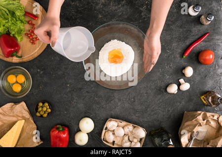 Vista parziale di donna versando acqua nella tazza di farina con pizza ingredienti su sfondo grigio Foto Stock