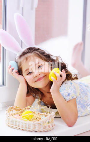 Ragazza con orecchie di coniglio con uova dipinte in mani sorridente sullo sfondo del cesto di pasqua seduto vicino a finestra Foto Stock