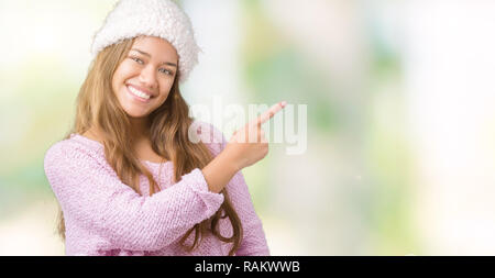 Giovane bella bruna donna che indossa un maglione e Cappello invernale su sfondo isolato allegro con un sorriso della faccia rivolta con la mano e il dito verso l'alto Foto Stock