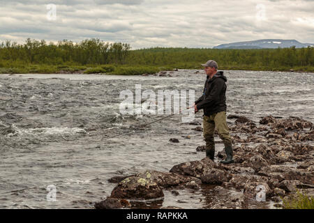 Uomo bianco, pesca in piedi sulle rocce della pesca, cercando felice e sorridente, mountain in background, fiume Rautas, Kiruna county, Lapponia svedese, Svezia Foto Stock