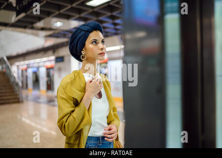 Una giovane e attraente donna musulmana in un velo e elegante color pastello vestiti è la consultazione di una mappa in una stazione ferroviaria per raggiungere la sua destinazione. Foto Stock