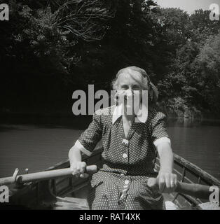 Degli anni Cinquanta, storico, un anziano ma energica signora indossando un macchiato o polka dot dress remare una barca di legno sulle rive di un fiume, Inghilterra, Regno Unito. Foto Stock