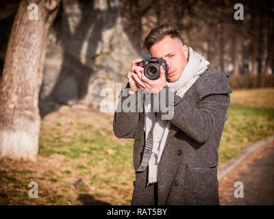 Bel giovane fotografo maschio tenendo fotografia professionale con fotocamera, all' aperto nel parco della città Foto Stock