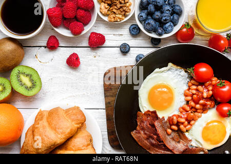 Colazione con caffè, uova fritte, pancetta, fagioli, cornetti, succo d'arancia con frutti e bacche. vista superiore Foto Stock