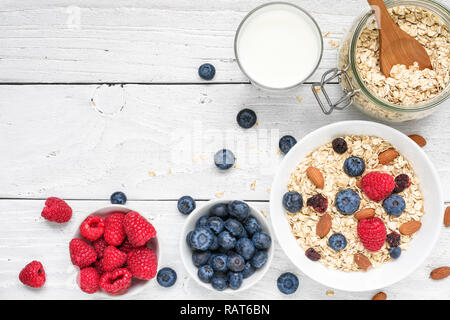 Sana colazione ingredienti. in casa di avena con lamponi e mirtilli, il latte e i dadi su bianco sullo sfondo di legno. vista superiore con spazio di copia Foto Stock