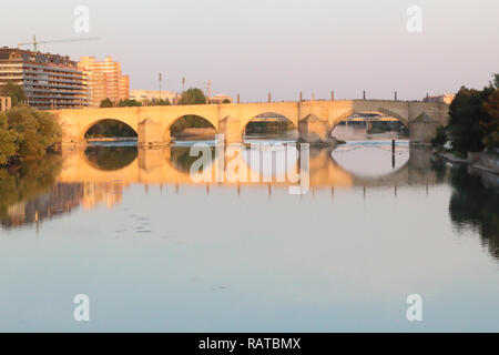Il Puente de Piedra ponte in pietra con archi a tutto sesto che riflette nel grande fiume Ebro acque al tramonto a Saragozza, regione di Aragona, Spagna Foto Stock