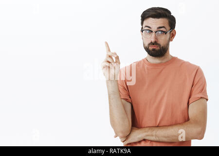 Simpatico insegnante con la barba in bicchieri sollevando il dito indice per fare il punto, insegnare qualcosa e dire cose intelligenti, in piedi saggio e intelligente su sfondo grigio parlando di studenti e noi oltre il muro grigio Foto Stock