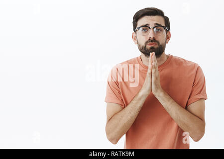 Vi preghiamo di Ho bisogno del tuo aiuto. Silly accattonaggio insicuro da stupidi guy in bicchieri scendere tenendo le mani nel pregare guardando con cute espressione triste in telecamera chiedendo a favore o il prestito di denaro in posa contro il muro grigio Foto Stock