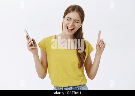 Affascinante carefree bella femmina in giallo t-shirt che indossa gli auricolari wireless dello smartphone di contenimento dancing gesticolando dalla gioia di chiudere gli occhi e cantare canzoni preferite su muro grigio Foto Stock