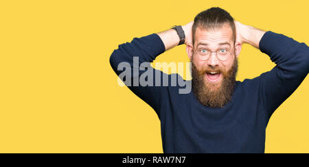 Giovane uomo biondo con gli occhiali e turtleneck maglione Crazy e spaventato con le mani sul capo, paura e sorpresa di shock con bocca aperta Foto Stock