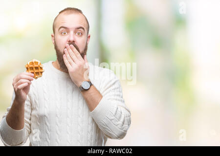 Giovani caucasici uomo parigamba mangiare dolci waffle su sfondo isolato coprono la bocca con la mano sconvolto con vergogna per errore, espressione di paura, sca Foto Stock