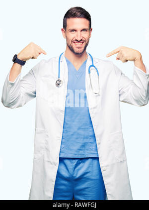 Bello medico uomo che indossa medical uniforme su sfondo isolato guardando fiduciosi con il sorriso sul volto, rivolto a se stessi con le dita in fieri e hap Foto Stock