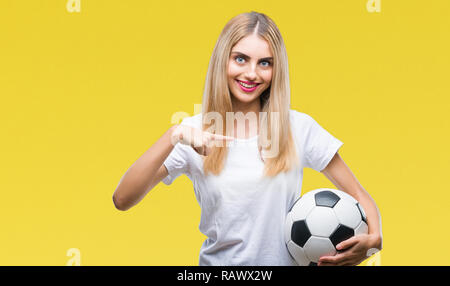 Giovane bella donna bionda tenendo palla calcio su sfondo isolato con sorpresa faccia puntare il dito a se stesso Foto Stock