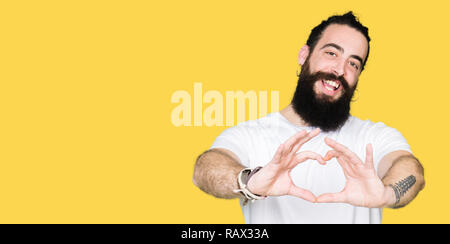 Tanga giovane uomo con i capelli lunghi e la barba che indossa casual t-shirt bianco sorridente in amore che mostra il simbolo del cuore e la forma con le mani. Concetto romantico. Foto Stock