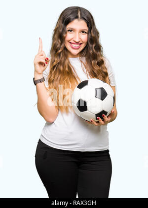 Giovane donna adulta holding soccer football ball sorpreso con un'idea o domanda puntare il dito con la faccia felice, numero uno Foto Stock