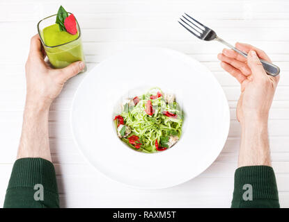 L'uomo azienda frullato verde e mangiare materie vegane spaghetti di zucchine, pomodori secchi e un dado formaggio su sfondo bianco in ristorante Foto Stock