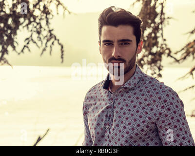 Ritratto di vita contemplativa bello dai capelli marrone giovane, con barba corta, indossa una maglietta accanto al pittoresco fiume o lago, guardando la telecamera a un si Foto Stock
