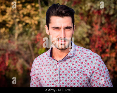 Ritratto di vita contemplativa bello dai capelli marrone giovane, con barba corta, indossa una maglietta accanto al pittoresco fiume o lago, guardando la telecamera a un si Foto Stock