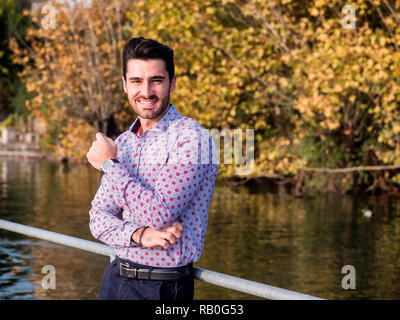 Ritratto di vita contemplativa bello dai capelli marrone giovane, con barba corta, indossa una maglietta accanto al pittoresco fiume o lago, guardando la telecamera a un si Foto Stock