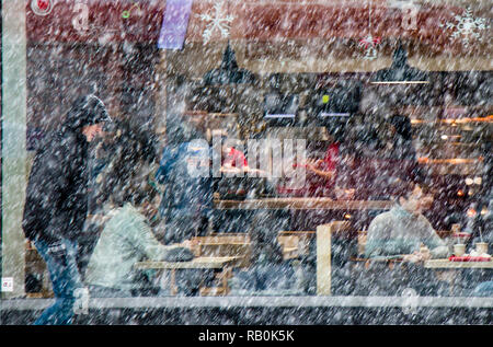 A Belgrado, in Serbia - Dicembre 15, 2018: sfocata uomo a camminare da sola strada di città in forte nevicata e la gente seduta in un ristorante finestra dietro di lui Foto Stock