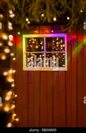 Coloratissima favola di Natale luci di notte si riflette in vetro antico dei riquadri della finestra rustico sul lato di un dipinto di rosso cabina di bosco. Foto Stock