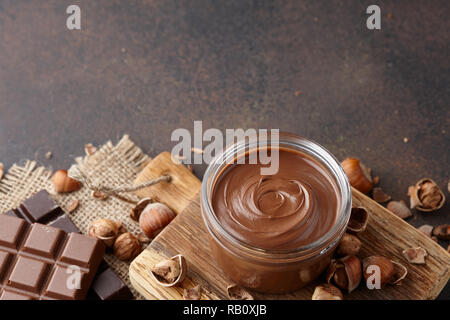 La diffusione di cioccolato o crema al torrone con nocciole in un barattolo di vetro, dadi e barrette di cioccolato sul bancone marrone Foto Stock